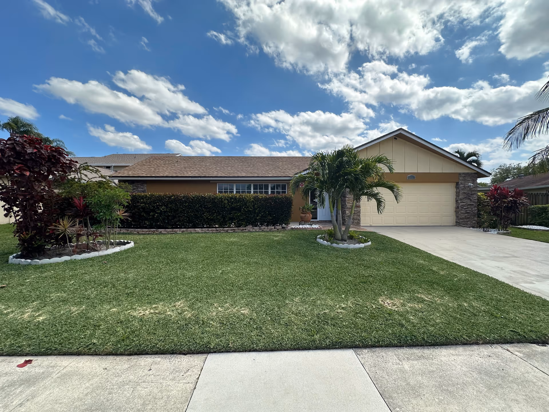 Residential front exterior with lawn, palm trees, and a two-car garage under sunny sky.