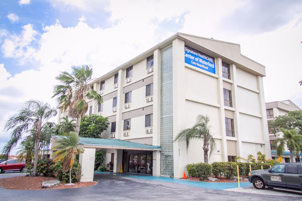 Three-story beige nursing home exterior with entrance, palm trees, and a parked vehicle.