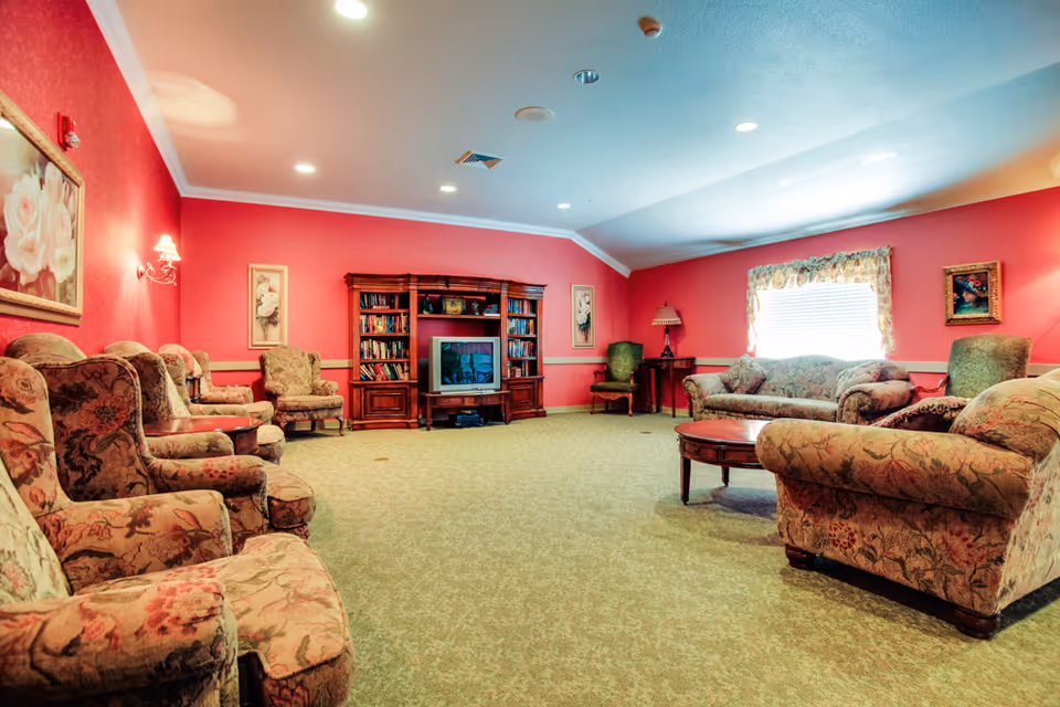 A spacious living room with pink walls and floral-patterned upholstered armchairs and sofas arranged around a wooden coffee table. A large wooden entertainment center with a TV and bookshelves is against the far wall. The room is carpeted and decorated with framed floral artwork and a window with floral curtains letting in natural light.