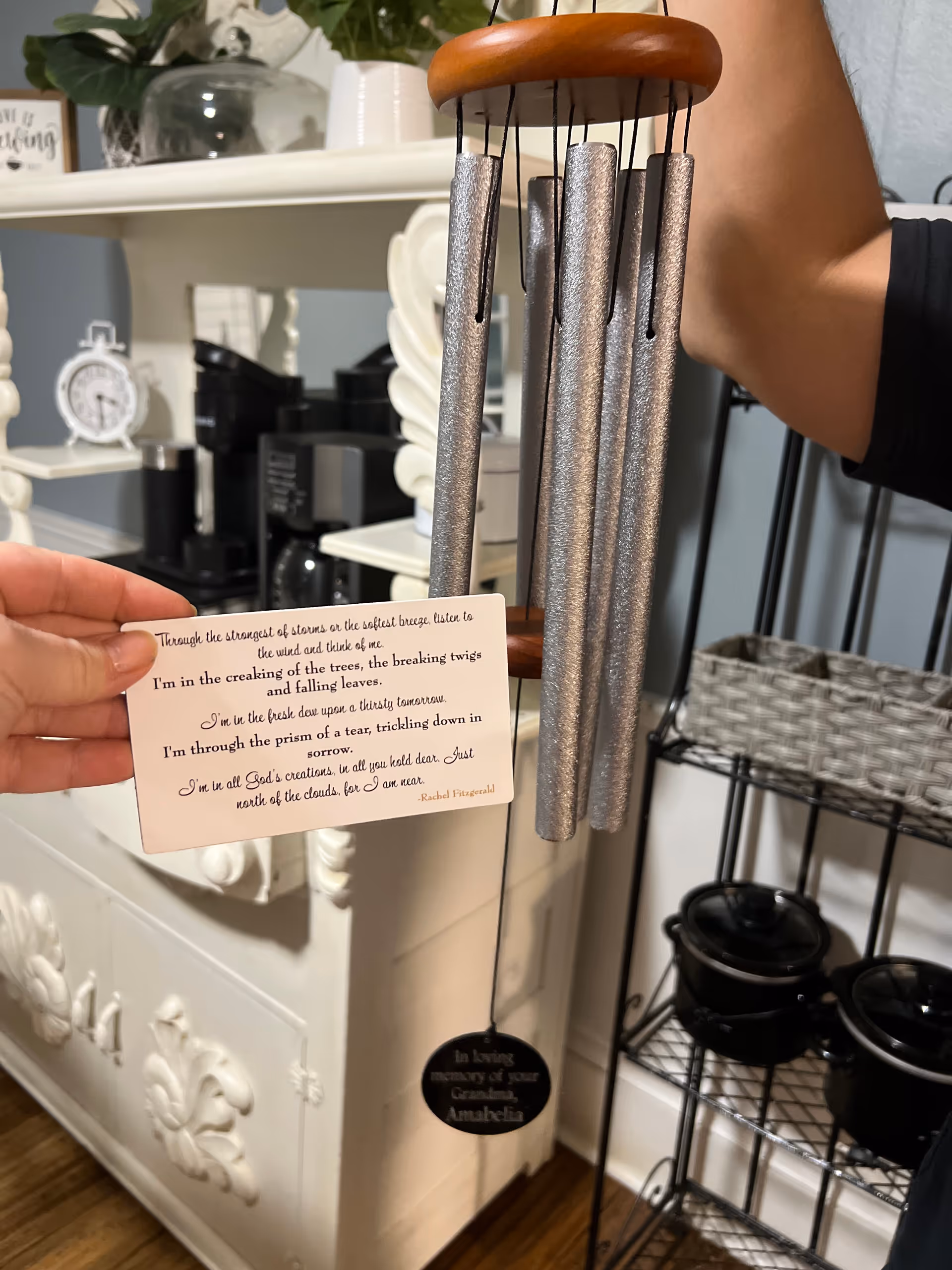 A hand holds a small poem card beside metallic wind chimes and decorative white furniture.