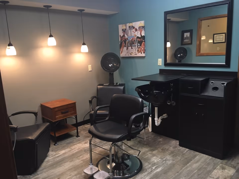 Interior of a hair salon area with a black salon chair in front of a black sink and counter. There are two black chairs, a small wooden side table, three hanging pendant lights, a large mirror on the wall, and a colorful painting of people sitting under hair dryers.