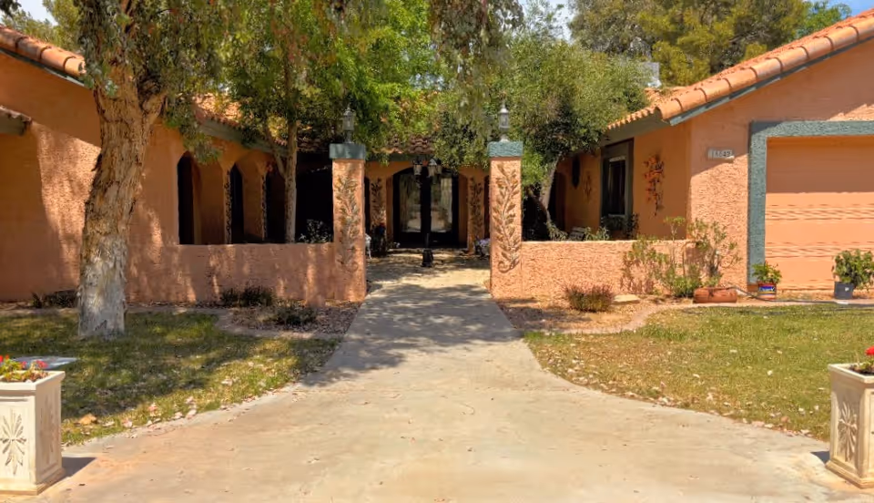 Front exterior view of a single-story building with peach-colored stucco walls and a tiled roof. There is a concrete pathway leading to the entrance, flanked by two decorative pillars and trees providing shade. The area has some grass and potted plants near the building.