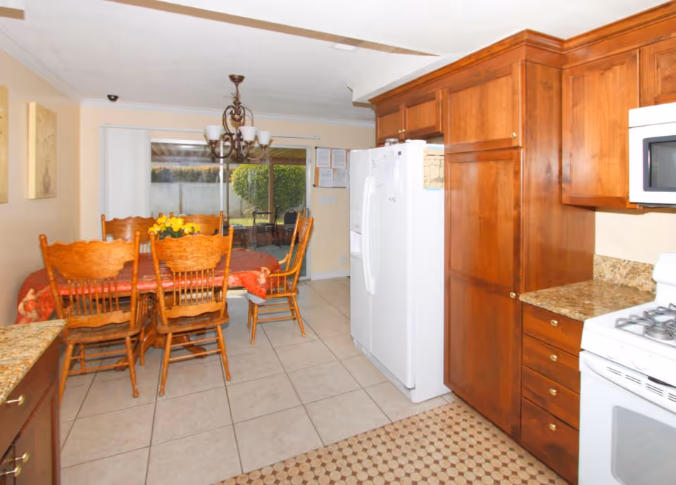 Wooden cabinets, appliances, and a dining table fill a warm kitchen area.