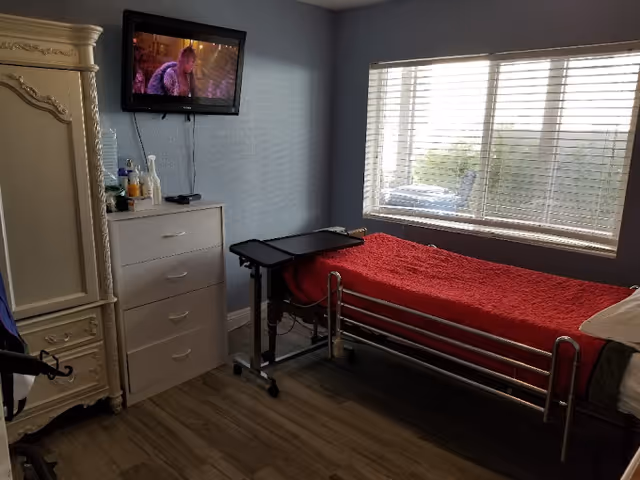A small bedroom with a single hospital-style bed covered with a red blanket, a white dresser with various items on top, a mounted flat-screen TV showing a scene from a movie, a large window with blinds letting in natural light, and a cream-colored ornate wardrobe.