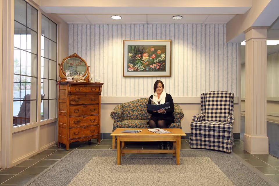 A woman sitting on a floral patterned couch in a cozy living room area, reading a book. The room features a wooden chest of drawers with a mirror, a checkered armchair, a wooden coffee table with magazines, and a framed floral painting on the wall. Large windows are on the left side, allowing natural light into the space.