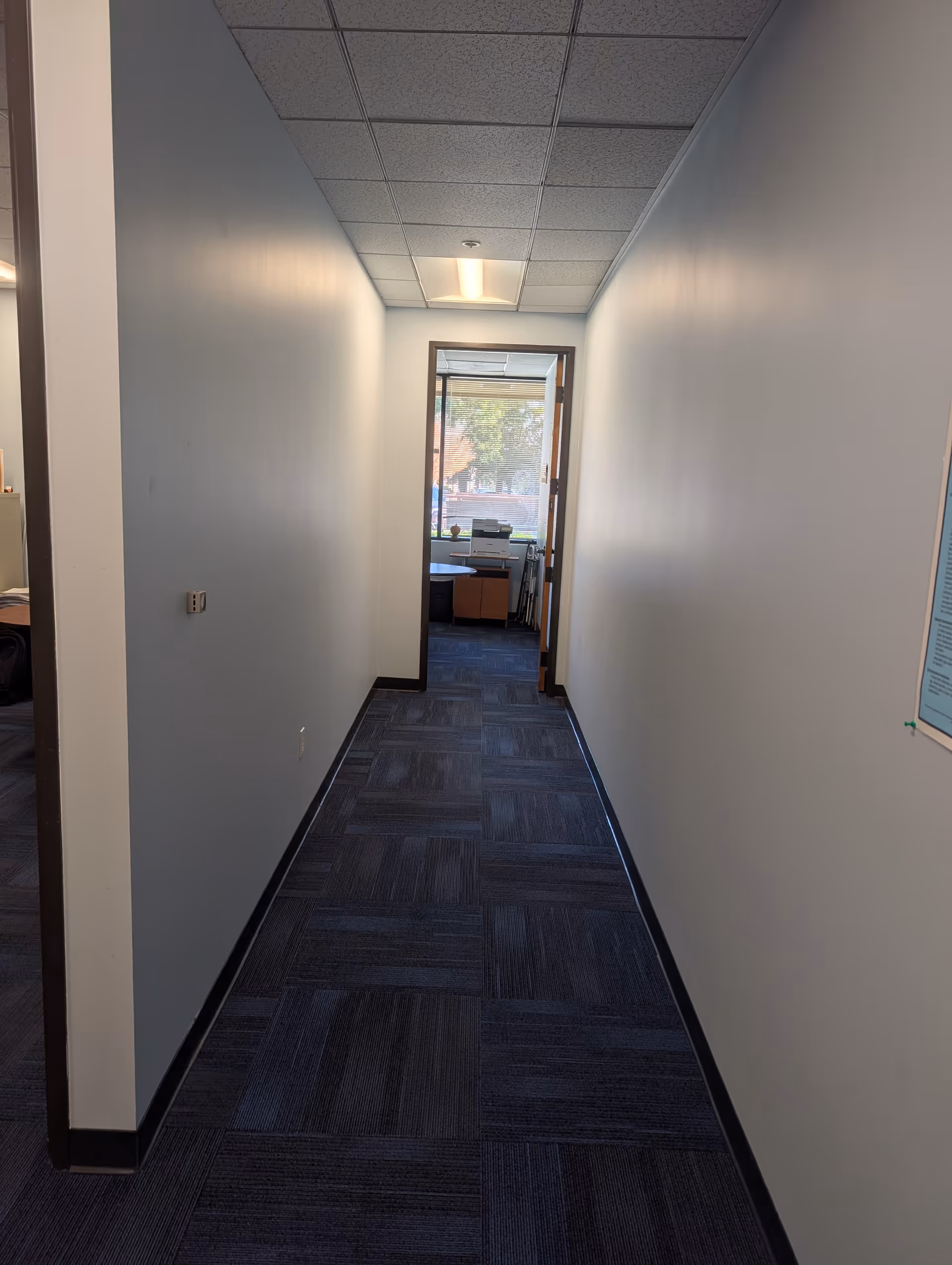 Narrow interior hallway with blue walls, carpet tiles, and a doorway at end.