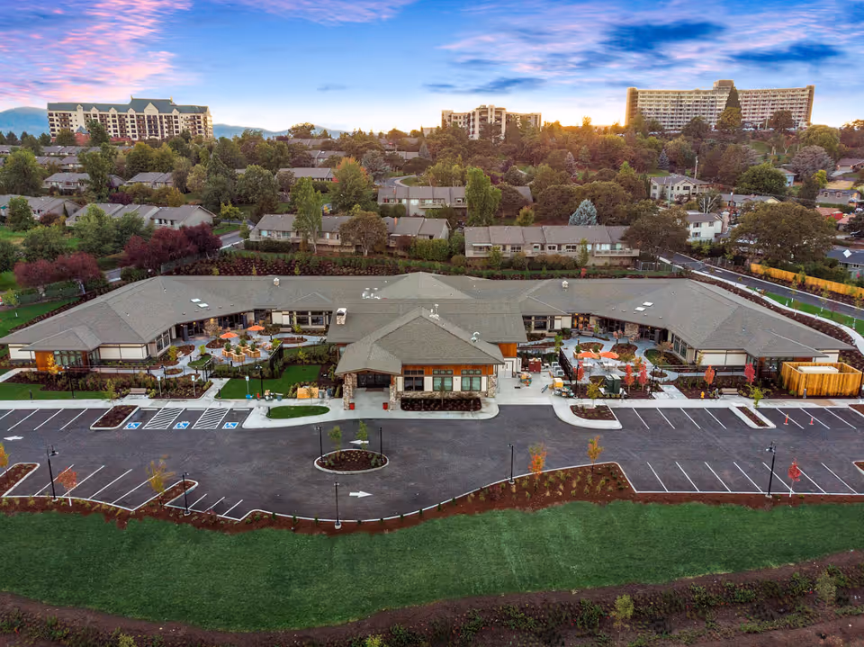 Aerial view of a single-story memory care building with patios, landscaping and a large parking lot set within a residential neighborhood at sunset.