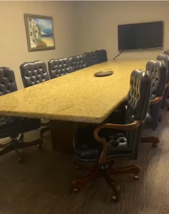 A long granite table with tufted leather chairs in a beige room with a wall-mounted TV.