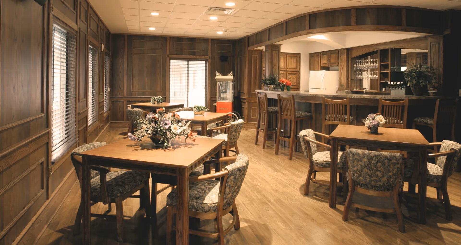 Wood-paneled communal dining area with wooden tables, upholstered chairs, and a bar/kitchenette in the back.