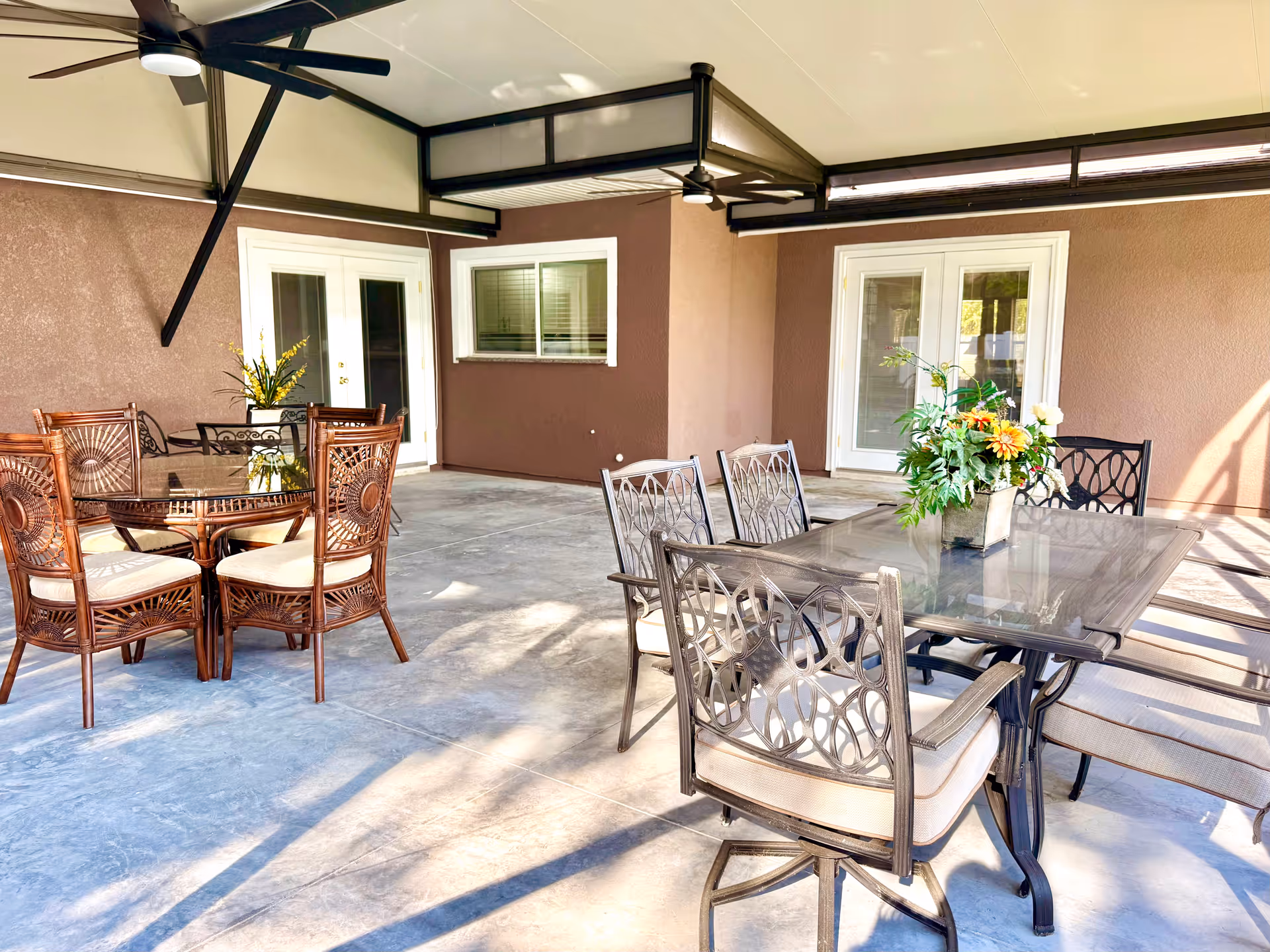 Covered outdoor patio with two dining sets, metal and wicker chairs, and potted plants.