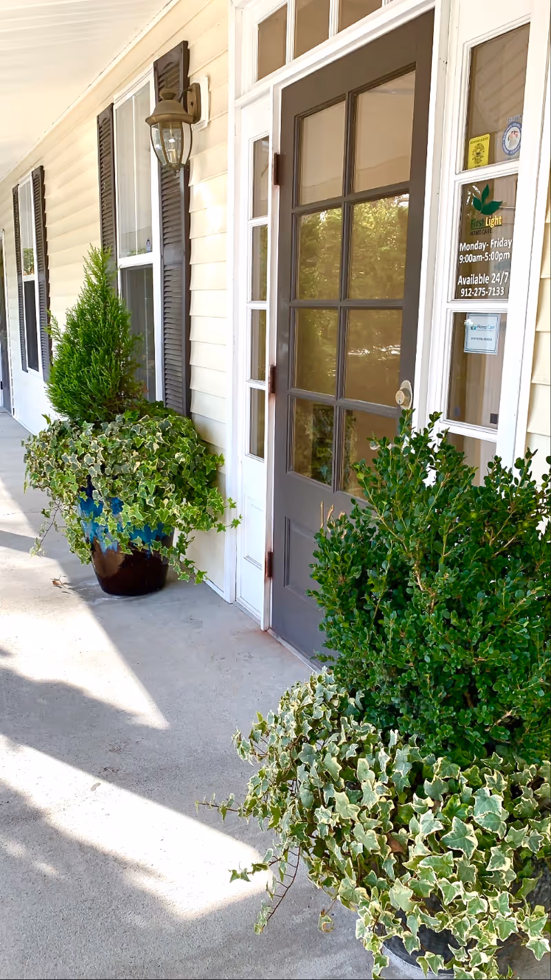 Exterior view of a FirstLight Home Care entrance with glass door, shutters, and potted plants.