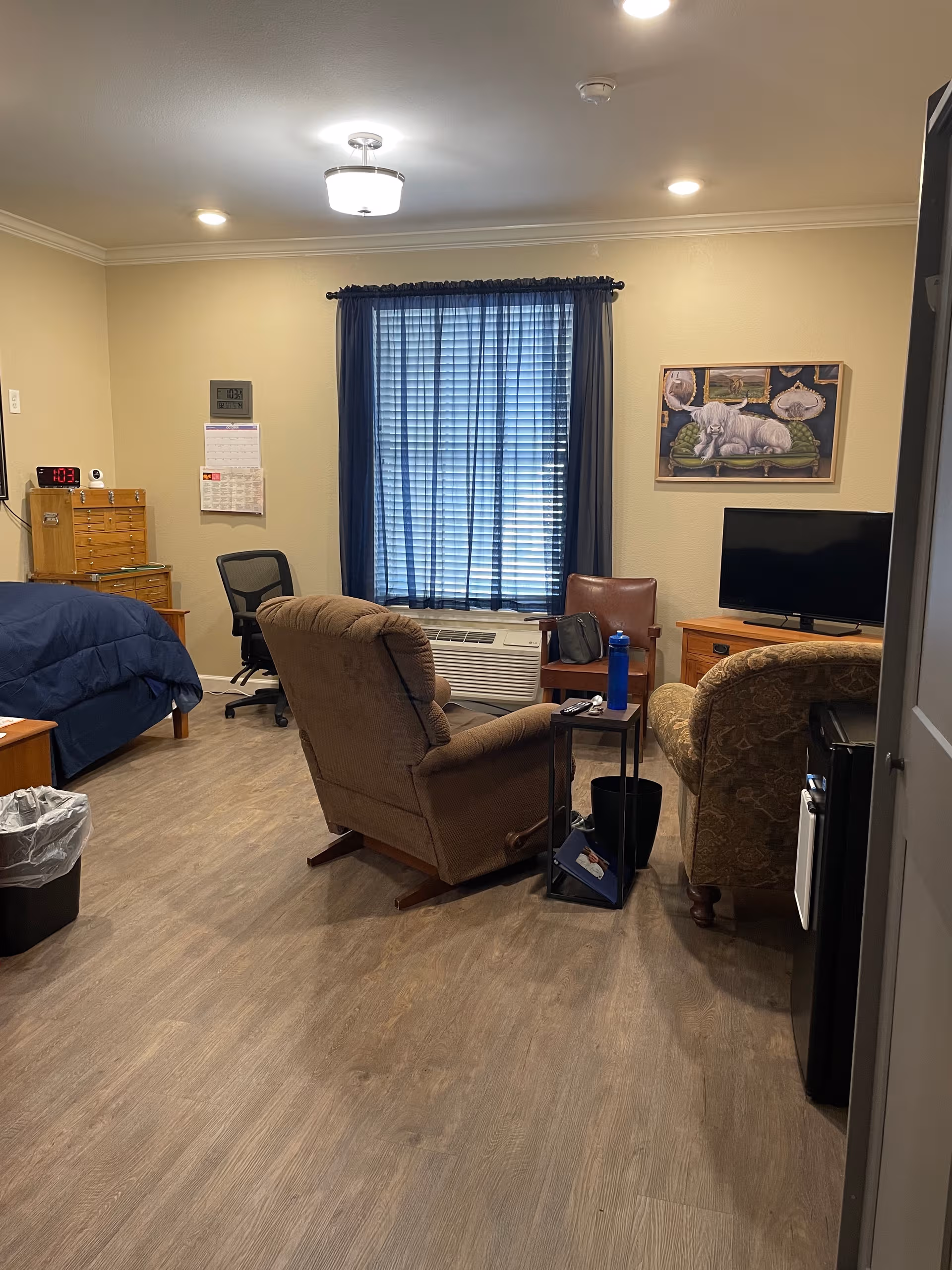 Cozy living room in a senior facility with seating, a TV, and a blue-curtained window.