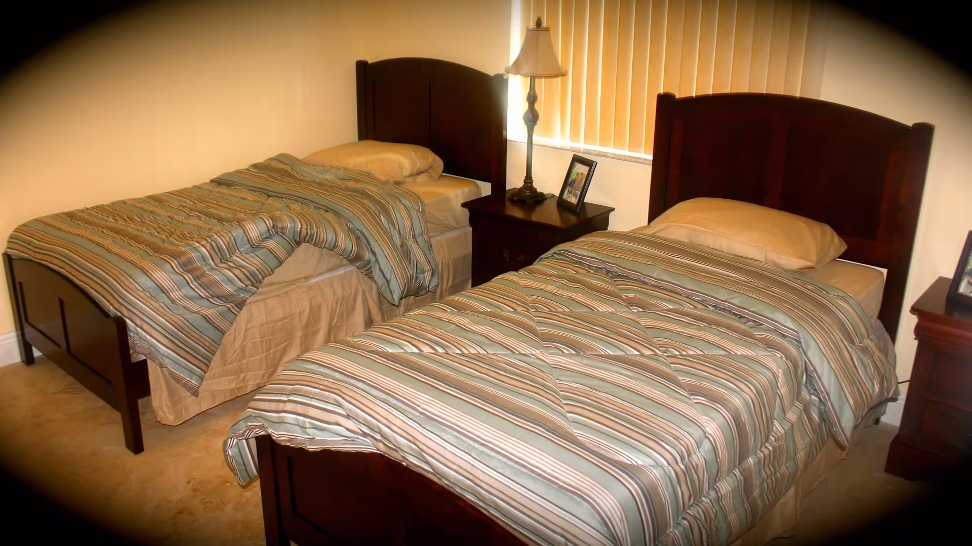 A bedroom with two single beds featuring dark wooden headboards and striped bedspreads in shades of blue, beige, and brown. Between the beds is a wooden nightstand with a lamp and a framed photo. Another framed photo is visible on a second nightstand to the right. The room has beige walls and a window with vertical blinds.