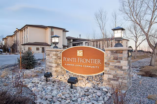 Entrance sign for Pointe Frontier, a senior living community, mounted on a stone base with two lantern-style lights on either side. Behind the sign, there are multi-story residential buildings and leafless trees under a clear sky.