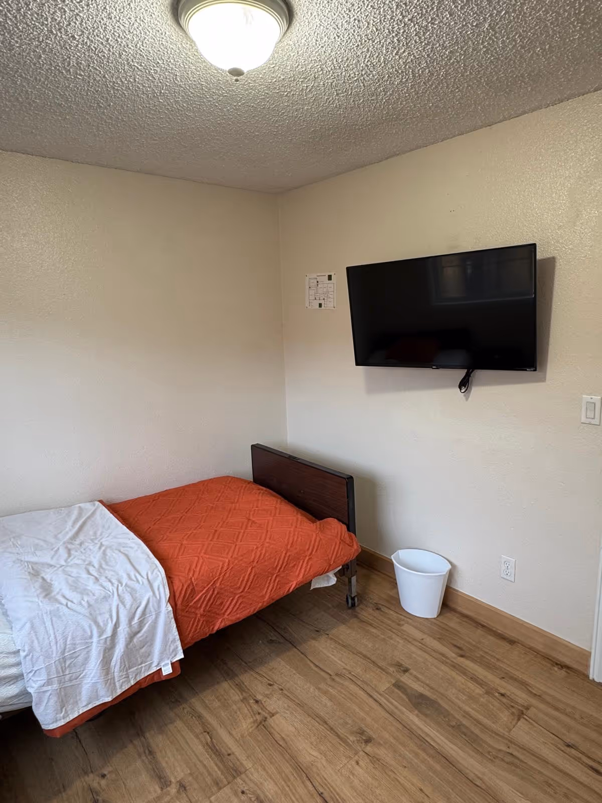 A small bedroom with a single bed, orange blanket, wall-mounted TV, and a white trash bin.