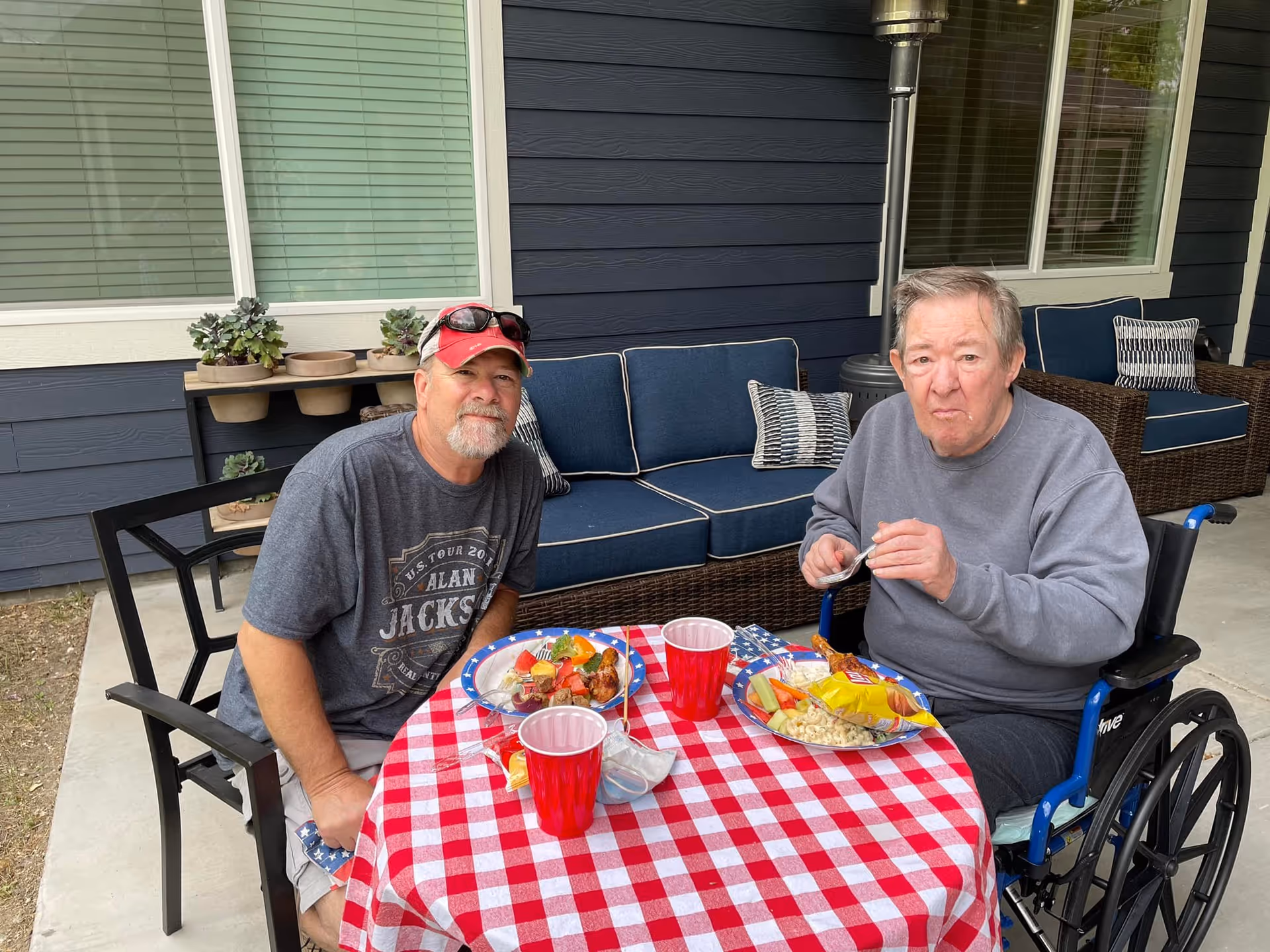 Two men sitting at a round table with a red and white checkered tablecloth outdoors. One man is seated in a wheelchair eating food from a plate, while the other man is sitting on a chair next to him. Behind them is a blue cushioned outdoor sofa and a small shelf with potted plants against a dark blue exterior wall with windows.