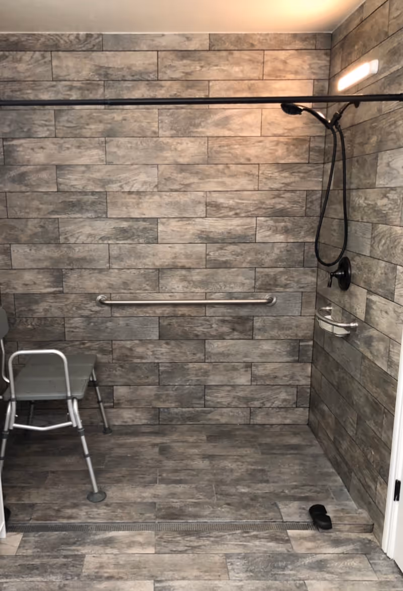 Accessible shower area with gray tile walls, grab bar, handheld shower, and chair.