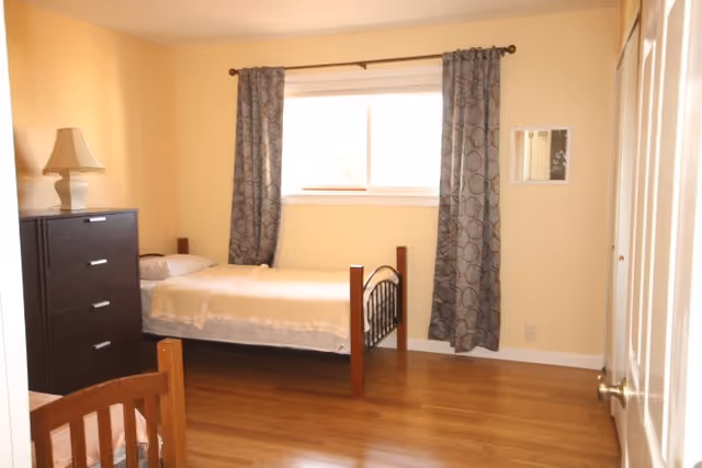 A simple bedroom with a single bed against the wall under a window with patterned curtains. There is a dark wooden dresser with a lamp on top next to the bed. The room has light yellow walls and wooden flooring.