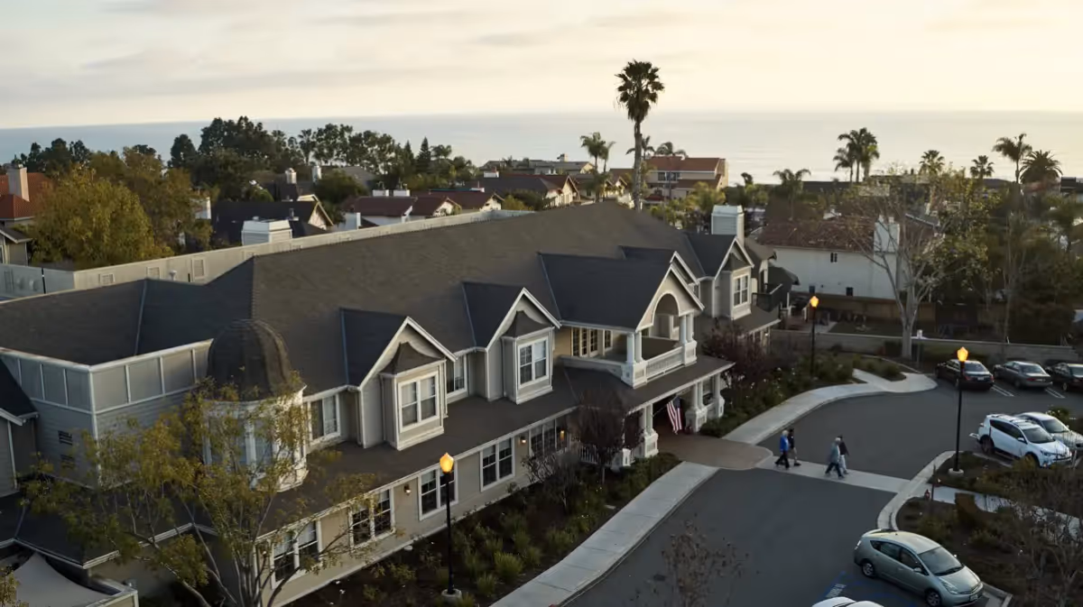 Aerial view of a large residential-style building with a dark roof and multiple dormer windows, surrounded by trees and a parking lot with several cars. Two people are walking on the driveway near the entrance. In the background, there are more houses, palm trees, and a distant view of the ocean under a cloudy sky.