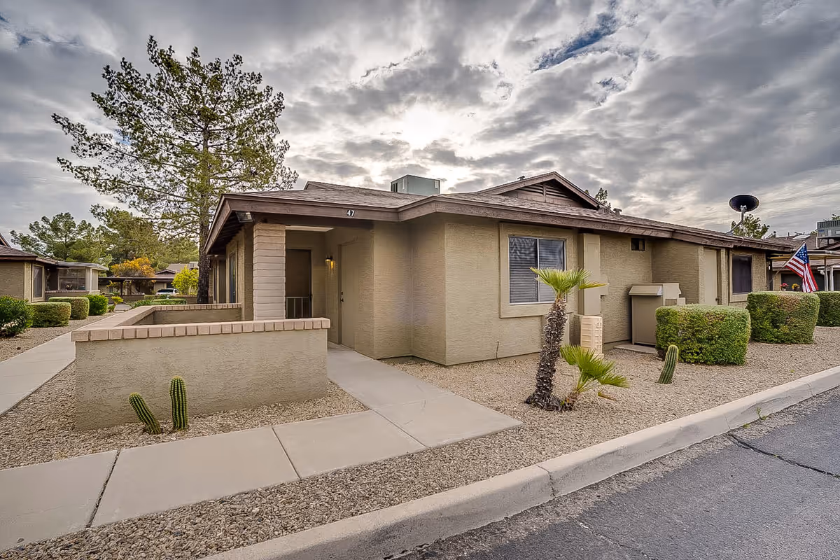 Single-story beige retirement home exterior with desert landscaping, sidewalk, and a flag.