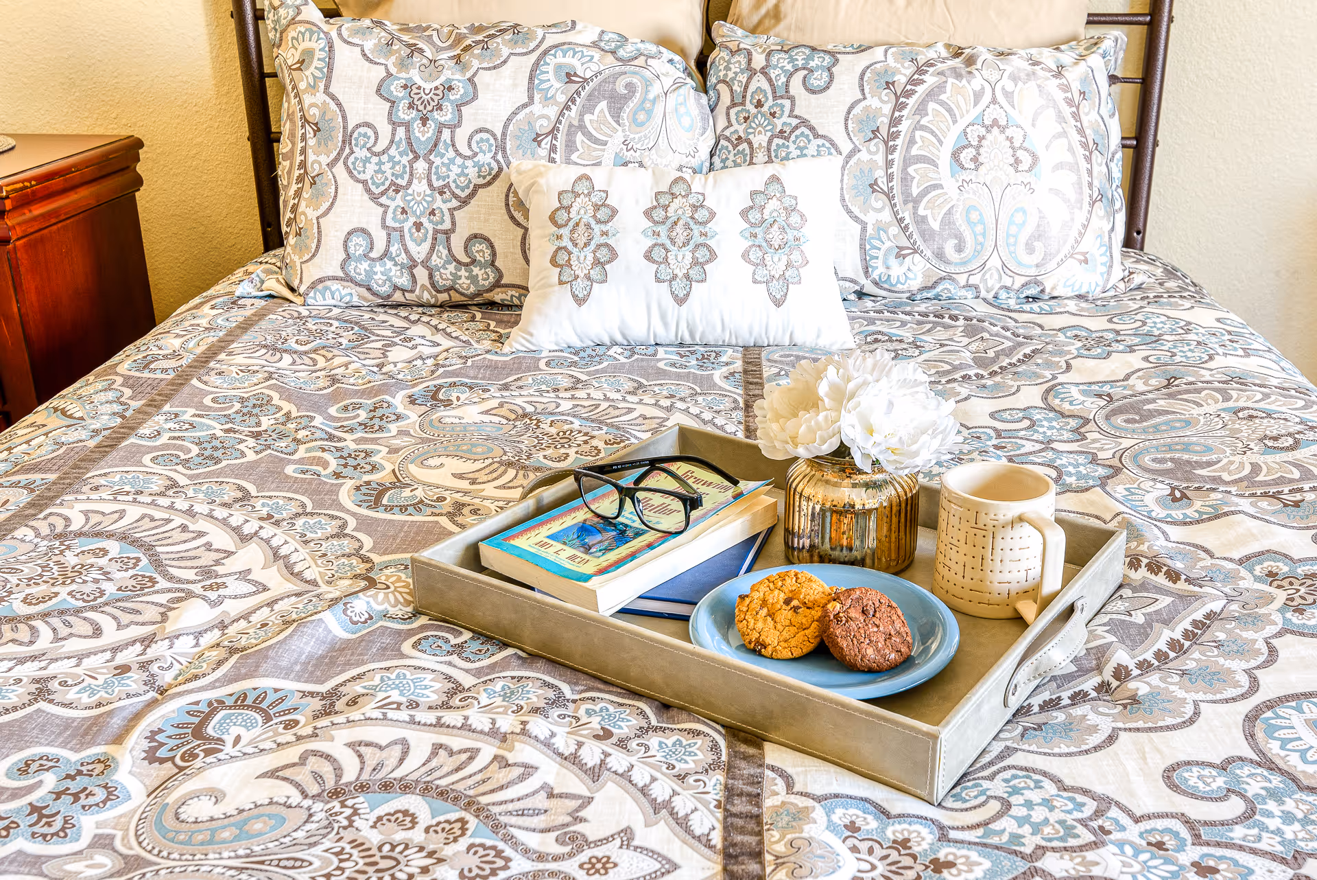 A neatly made bed with a tray of books, flowers, and cookies on the quilt.
