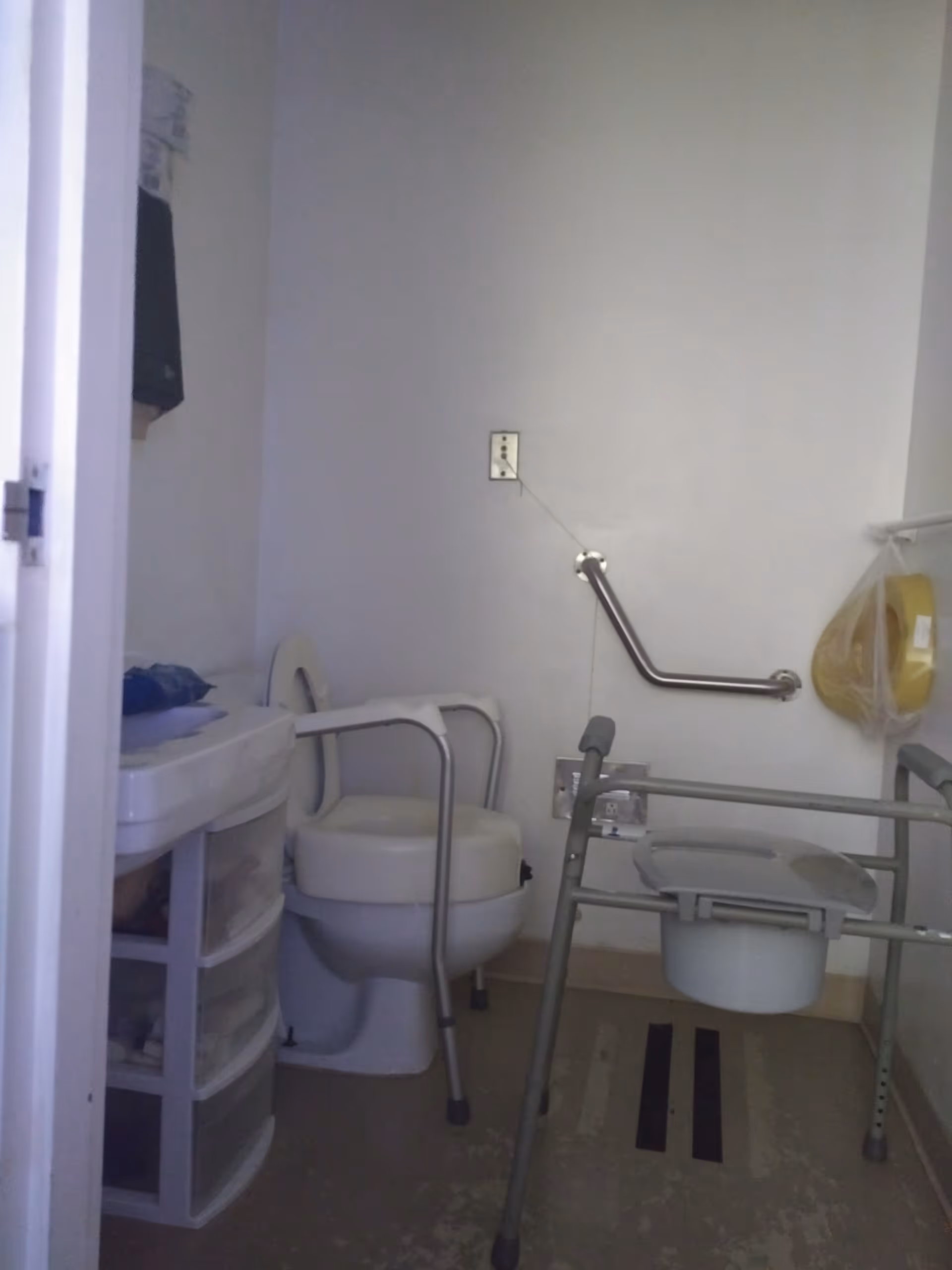 Small accessible bathroom with toilet, walker, grab bars, and sink.