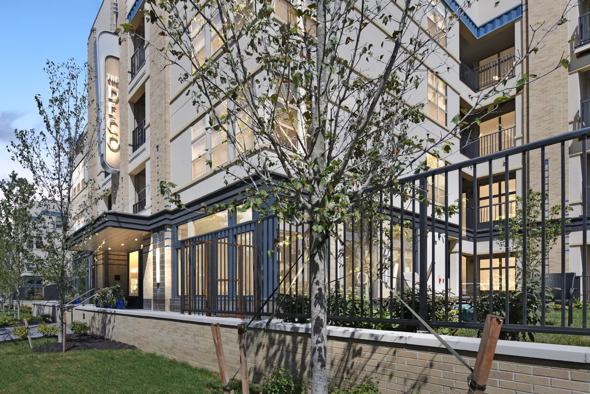 Exterior front entrance of The Deco senior living building with a lit vertical sign, balconies, fencing, and landscaped trees.