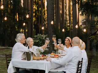 Six elderly people toast glasses around a white tablecloth table outdoors under string lights.