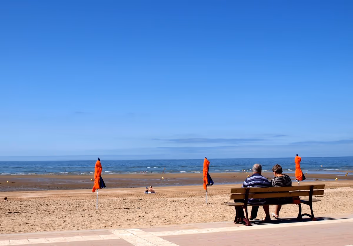 Two people sit on a bench along a sandy beach promenade with orange umbrellas.