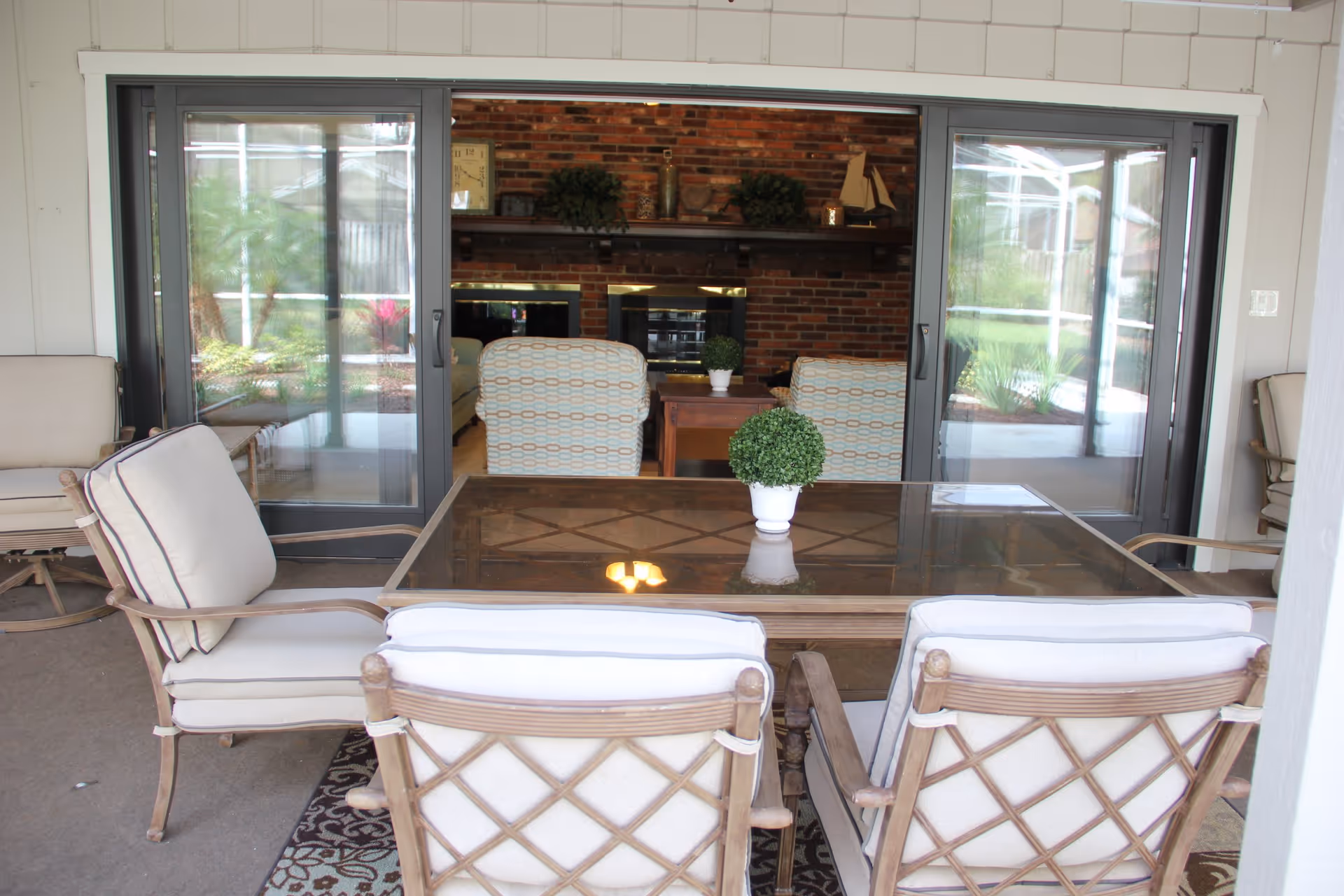 Outdoor patio area with cushioned chairs around a glass-top table, a small potted plant on the table, and sliding glass doors leading to an indoor living room with patterned armchairs and a brick fireplace.