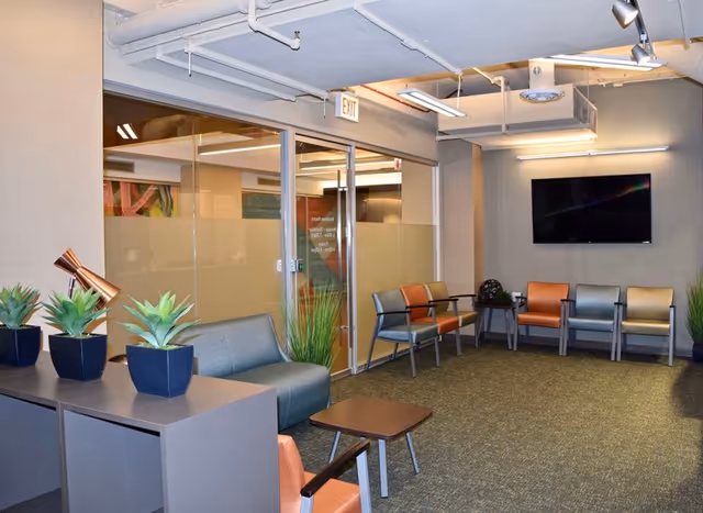 Modern interior waiting area with colorful chairs, glass walls, plants, and a wall-mounted TV.