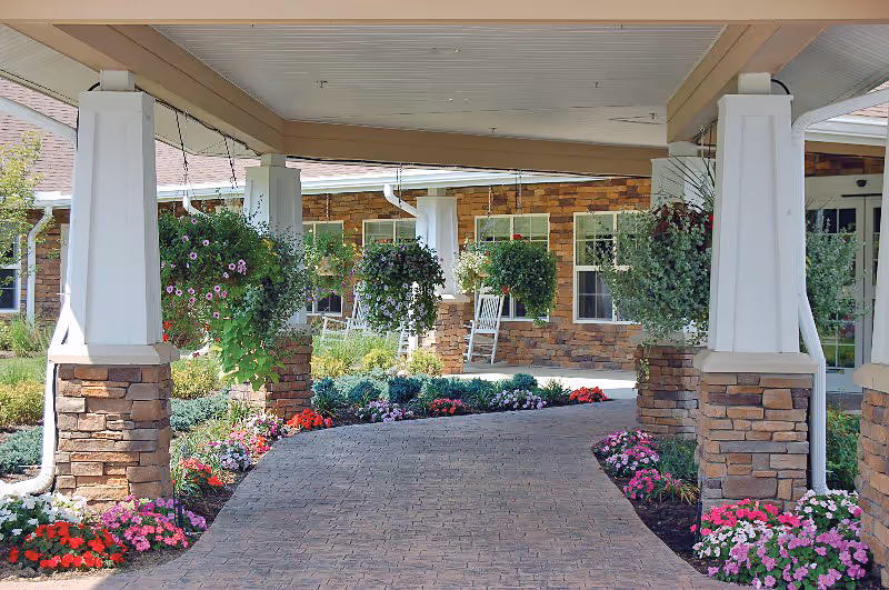 Covered entrance with stone pillars, hanging flower baskets, brick driveway and rocking chairs at the front of a senior living facility.