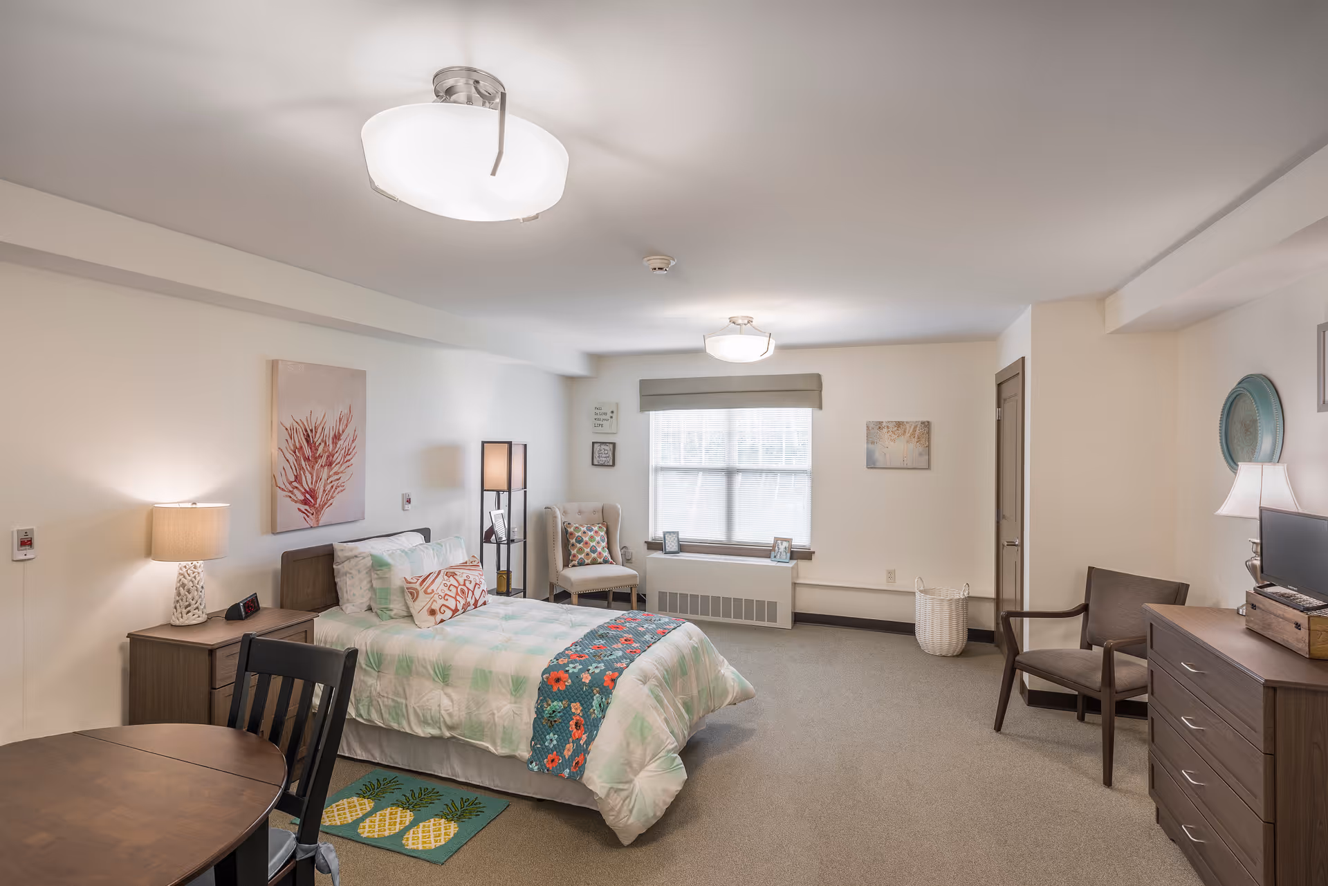 Well-lit, furnished senior bedroom with a bed, nightstand, dresser, seating area, and a window.