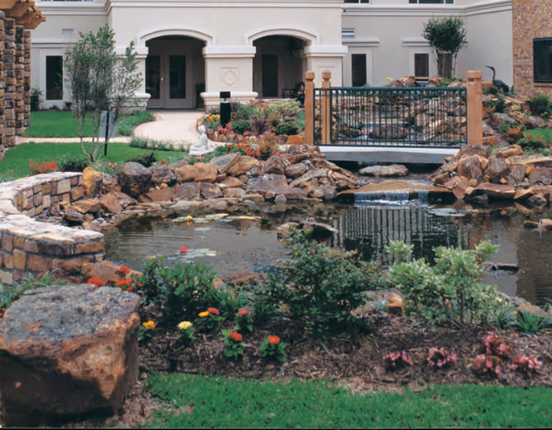 A landscaped outdoor garden area with a small pond surrounded by rocks and colorful flowers. There is a decorative bridge over the pond and a white statue near the pathway leading to a building with arched doorways and windows.