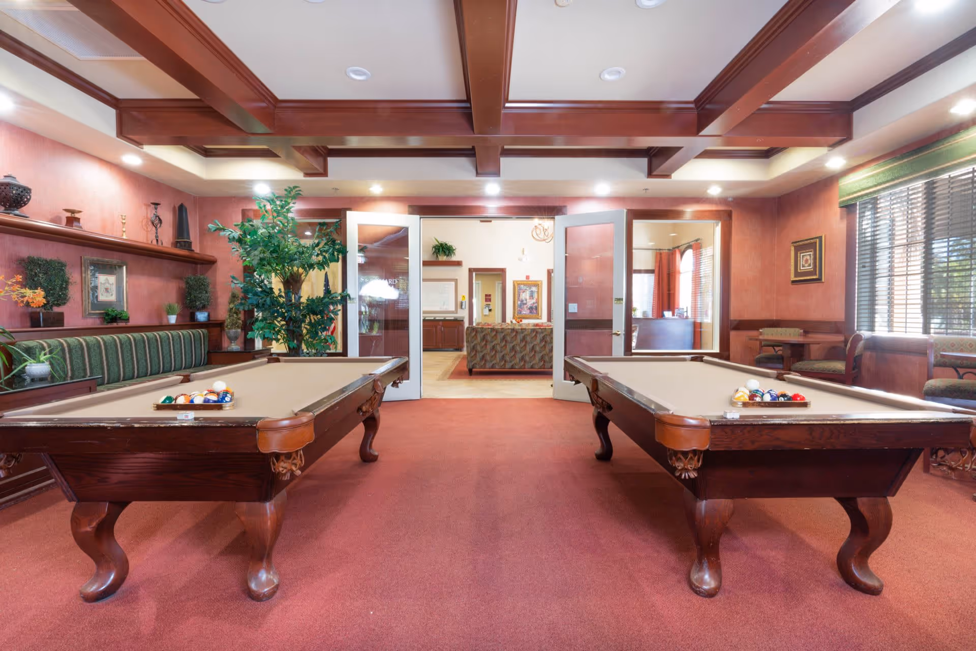 Interior view of a recreational room with two pool tables, a red carpeted floor, wooden ceiling beams, and a seating area with green striped cushions along the left wall. There are plants and decorative items on shelves, large windows with blinds on the right, and double glass doors leading to another room with more seating.