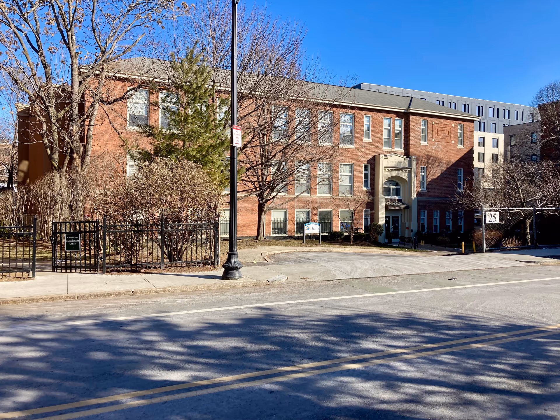 Three-story red brick assisted-living building with trees, a small fenced lawn and a street in front under a clear blue sky.