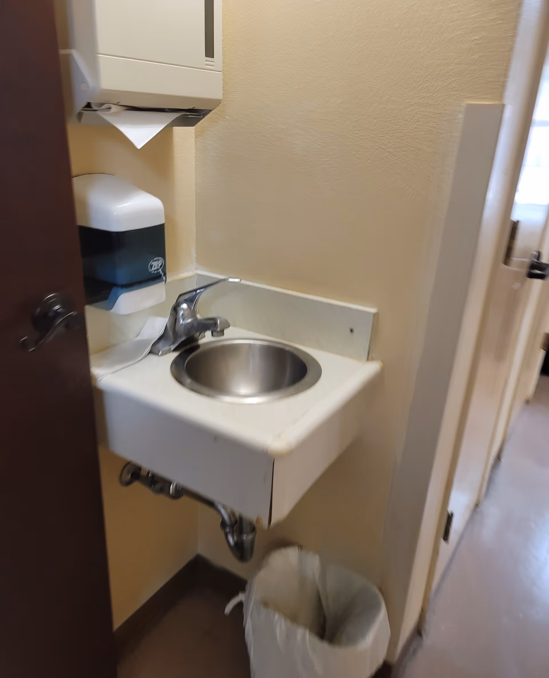 Small wall-mounted sink with faucet, paper towel dispenser, and trash can in a narrow hallway bathroom.
