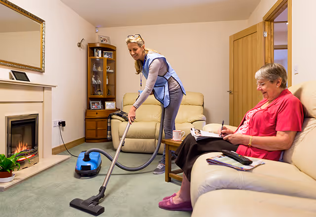 Caregiver vacuums the living room as a resident reads on a cream sofa.