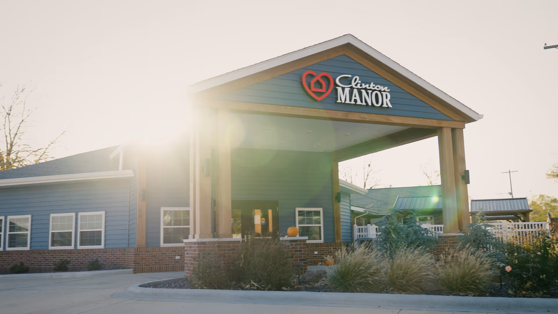 Exterior view of Clinton Manor Living Center building with a covered entrance, blue siding, brick accents, and landscaping in front. The sun is shining brightly, creating lens flare in the image.