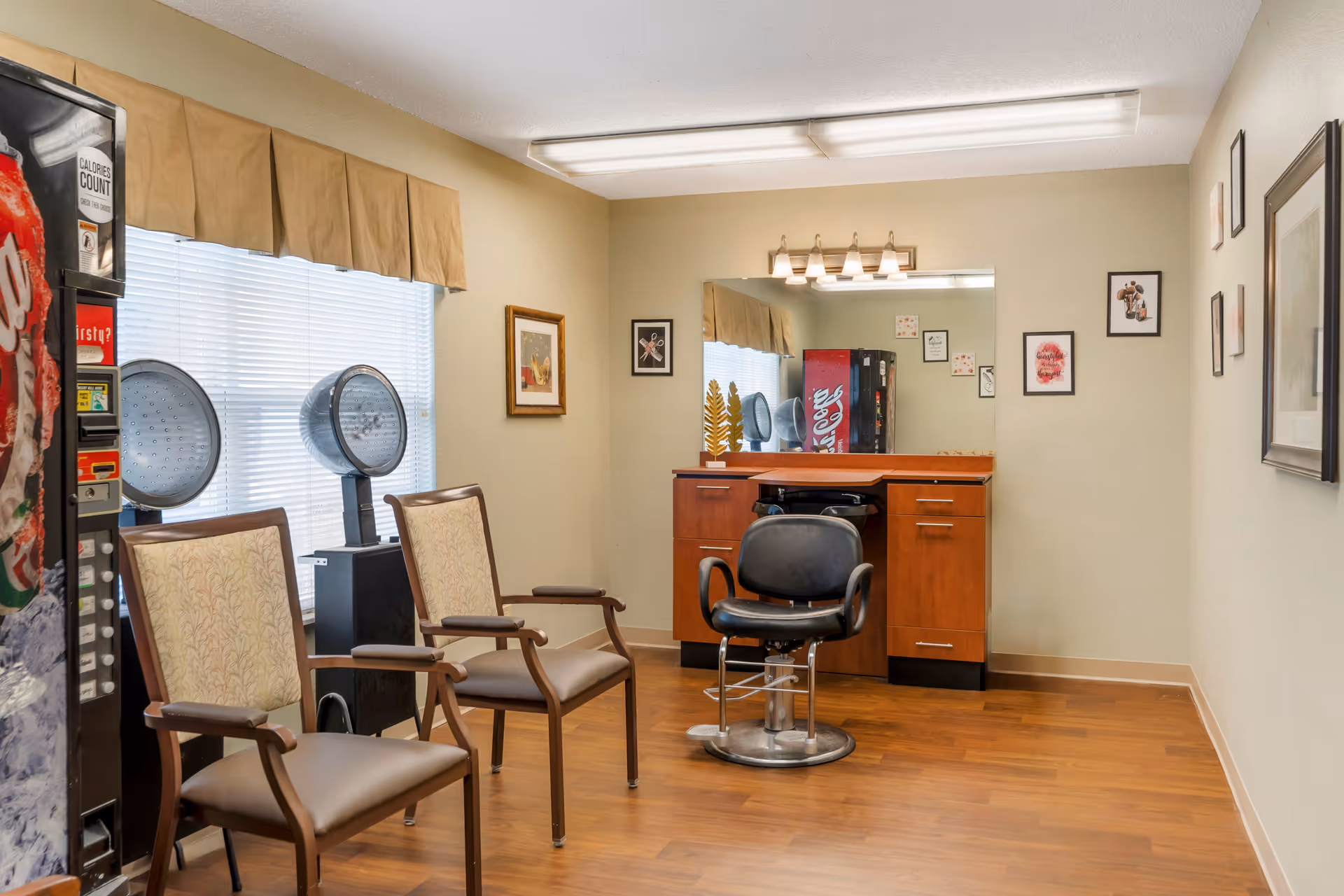 Small salon room with a styling chair in front of a mirror, two waiting chairs, hair dryers, and a soda vending machine.