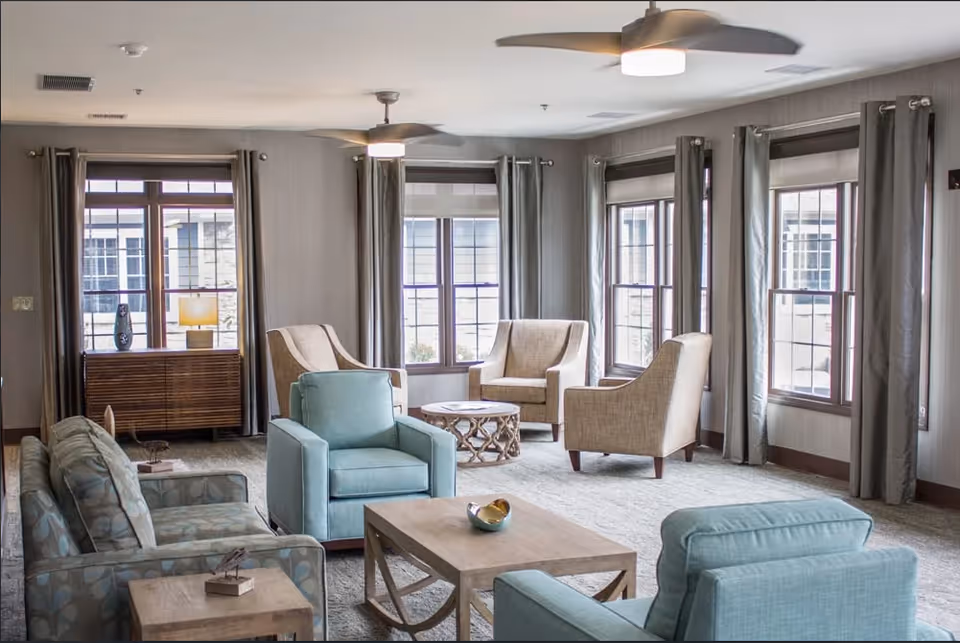 A bright and spacious living room with multiple large windows covered with gray curtains. The room features a mix of beige and light blue upholstered armchairs and sofas arranged around wooden coffee tables. Ceiling fans with lights hang from the ceiling, and a wooden sideboard with a lamp and decorative vase is positioned against one wall.