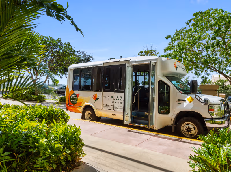 Shuttle bus branded The Plaza at Moanalua parked outside amid greenery.