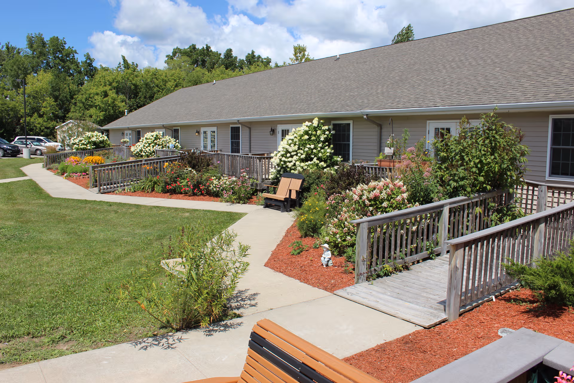 Single-story senior living building with wooden walkways, benches, and colorful gardens outside.