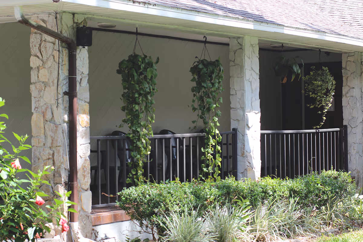 Covered porch with stone columns, black railing, hanging potted vines, chairs, and landscaping in front.