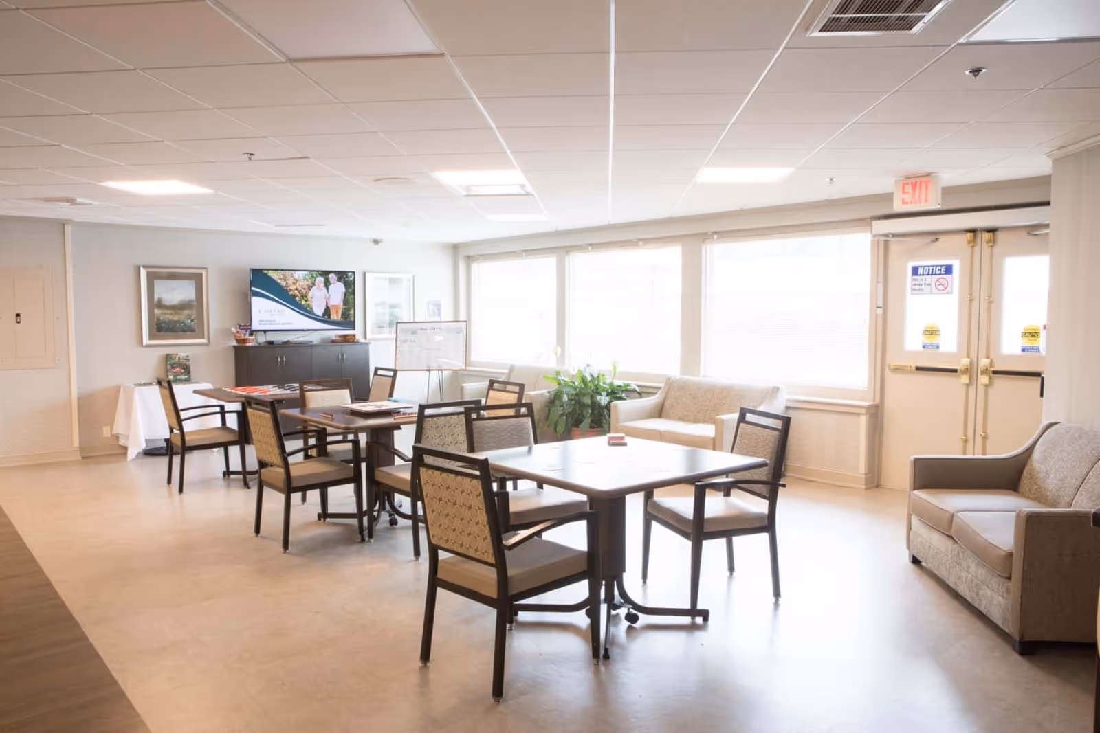 Bright communal dining/activity room with tables and chairs, sofas, a TV, and large windows letting in natural light.