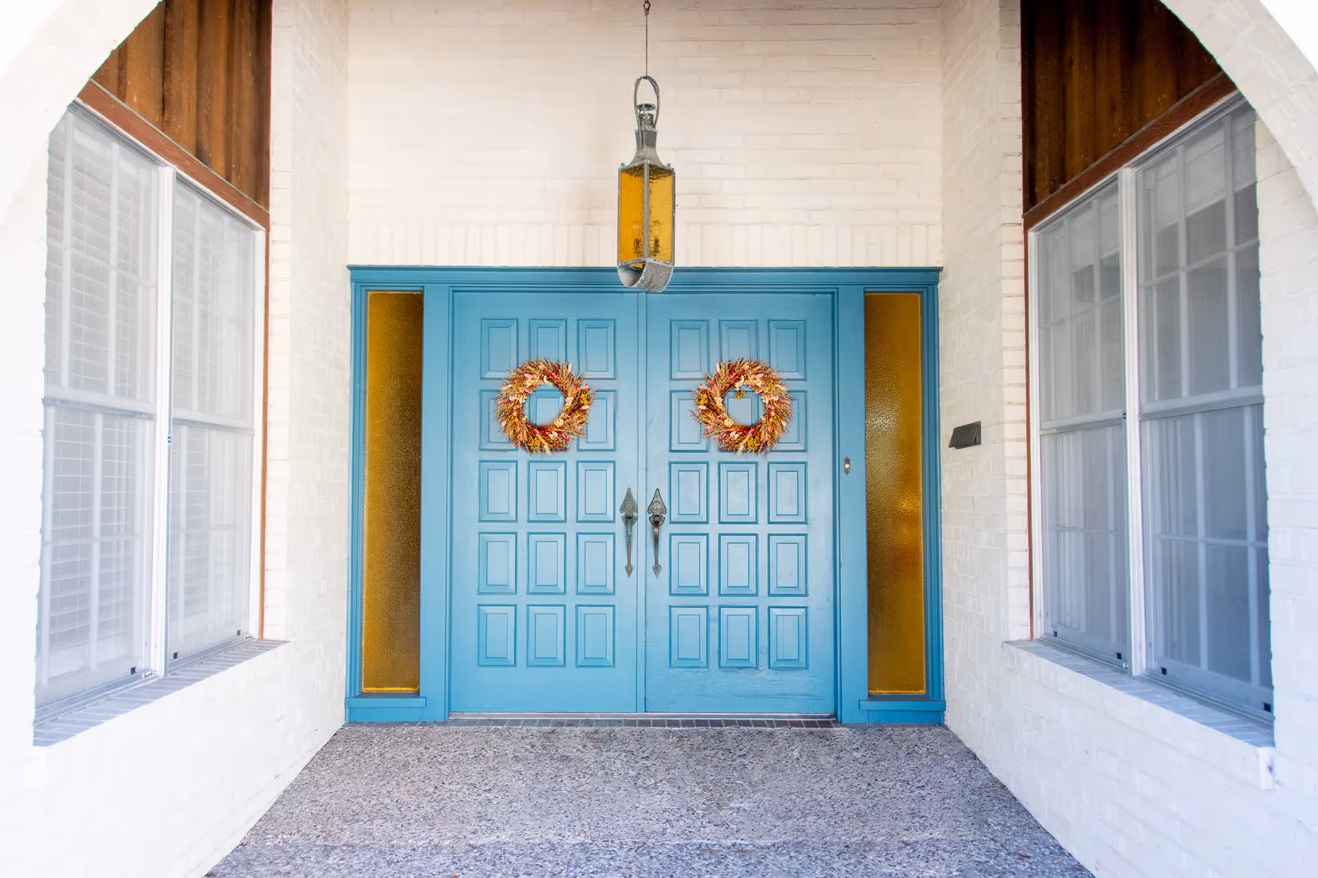 Front entrance of a building with double blue doors decorated with two autumn-themed wreaths. The doors are flanked by narrow amber-colored glass panels and white brick walls. There are windows on both sides and a hanging lantern light above the doors.