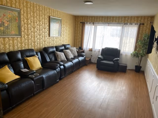 A living room with a row of black leather recliner chairs along the left wall, each with cushions in yellow and patterned designs. The room has wooden flooring, yellow patterned wallpaper, two framed floral paintings on the wall, a large window with sheer white curtains, a single black leather armchair near the window, a potted plant in the corner, and a flat-screen TV mounted on the right wall.