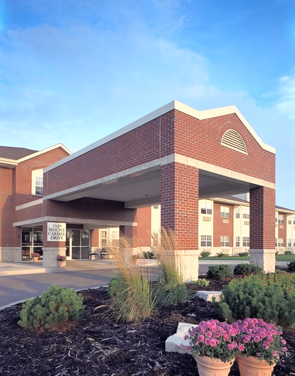 Front entrance of a brick senior living building with a covered porte-cochere, landscaped beds and potted flowers.