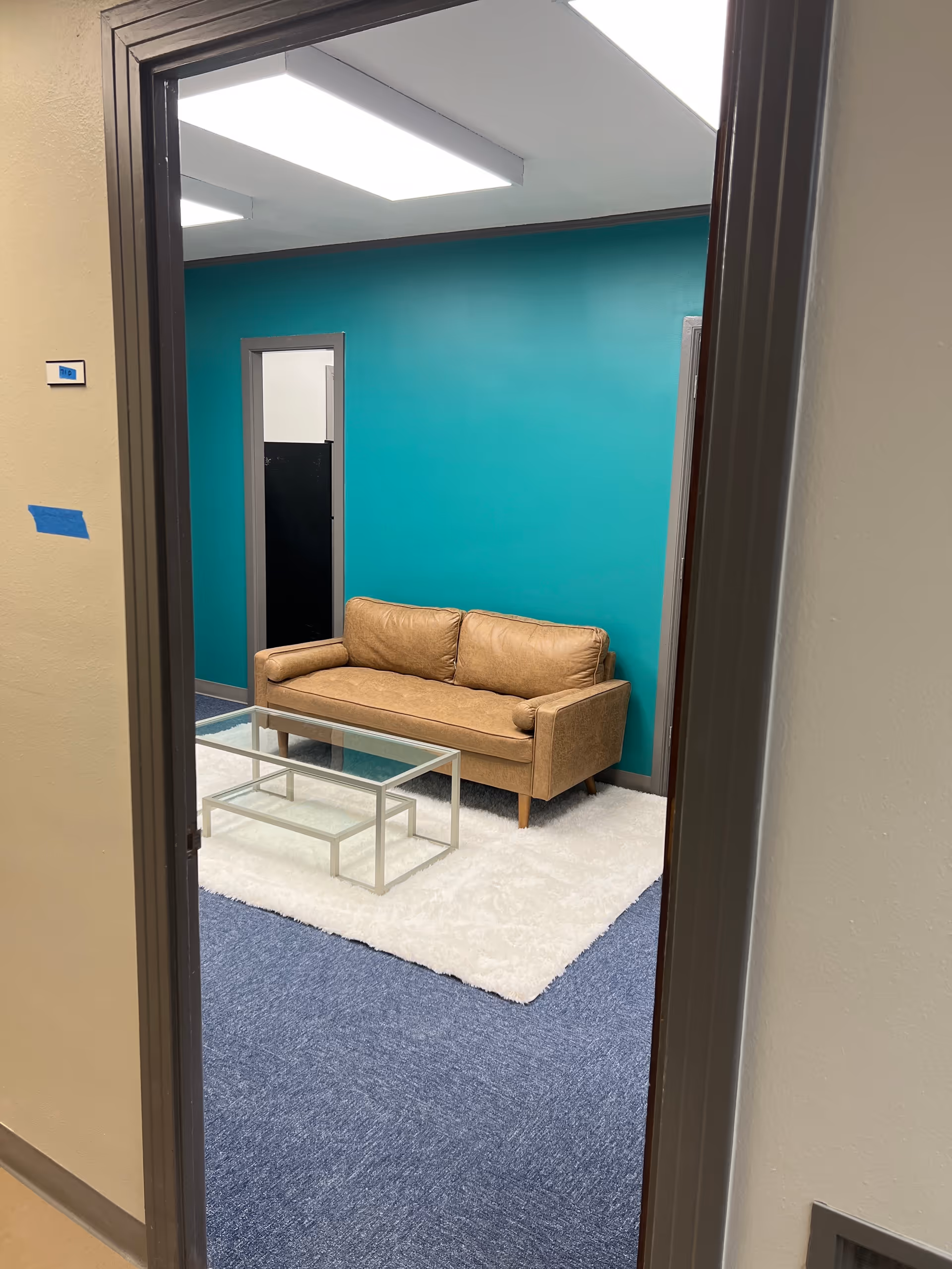 Teal-walled lounge with tan sofa, glass coffee table, white rug, and blue carpet.