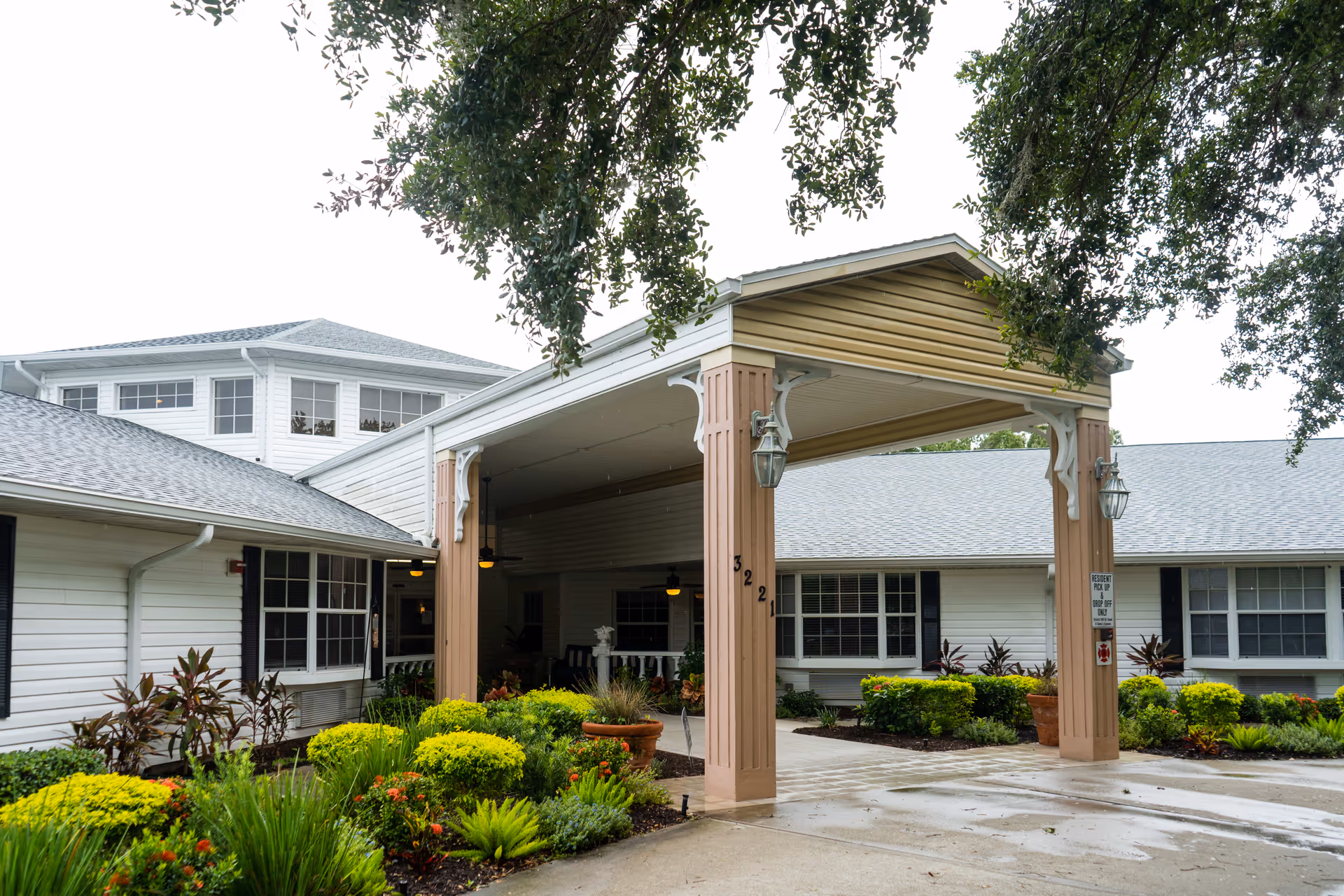 Entrance of a senior living facility named Heron House with a covered drop-off area supported by columns, surrounded by well-maintained landscaping including green shrubs and flowering plants. The building is white with multiple windows and a gray shingled roof, and there are ceiling fans under the covered area.